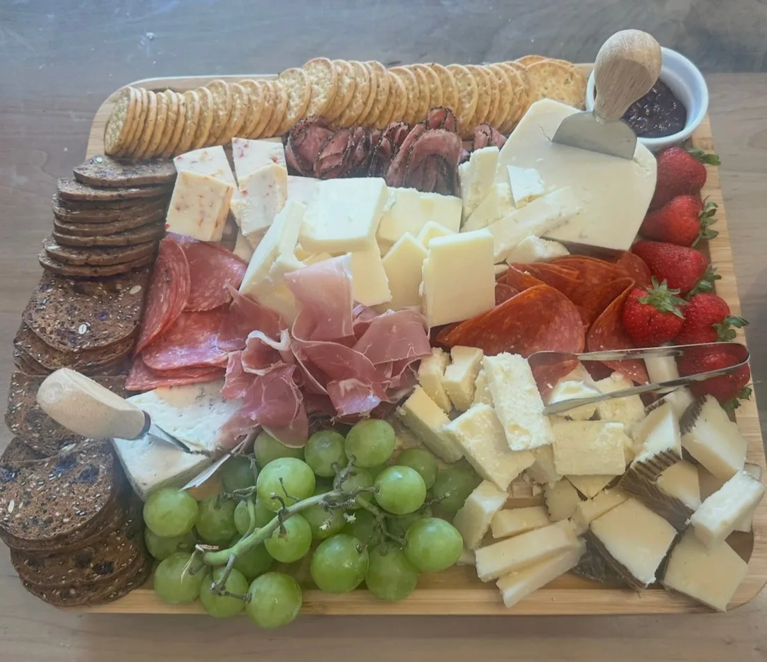 A board of cheeses and folded meats with green grapes, strawberries, crackers and preserves