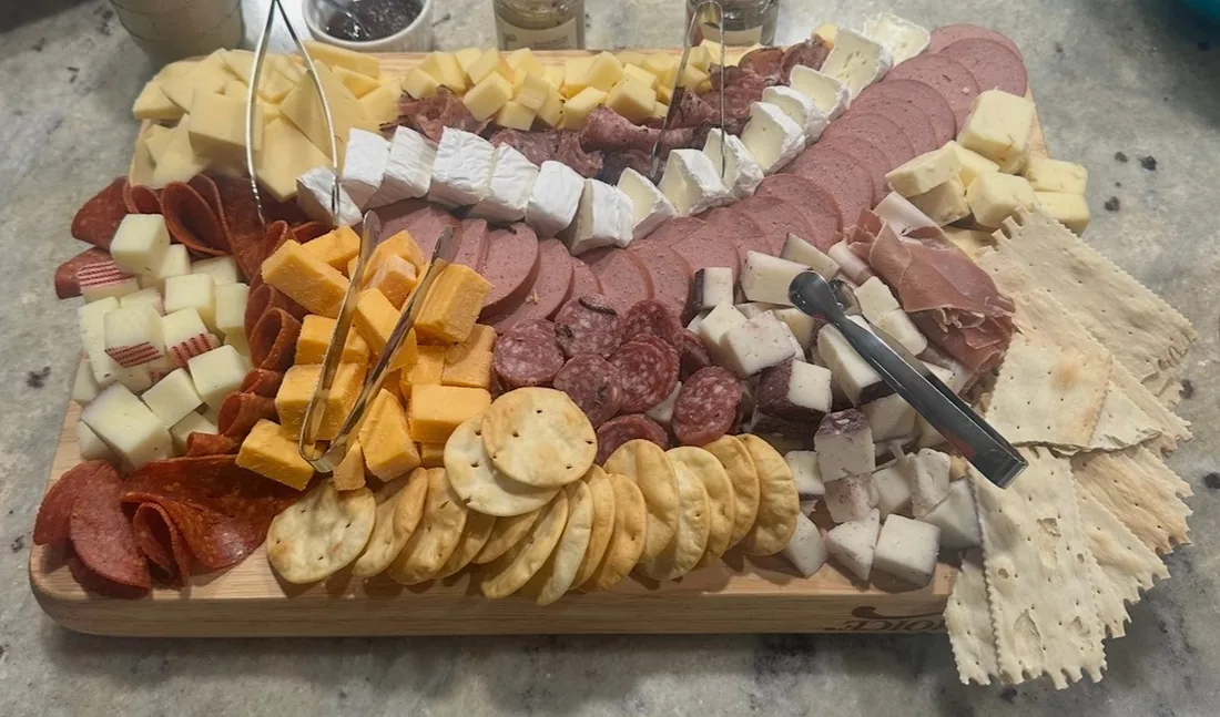 A rectangular wooden board arranged with sliced meats, brie, cubes of cheese and a row of crackers