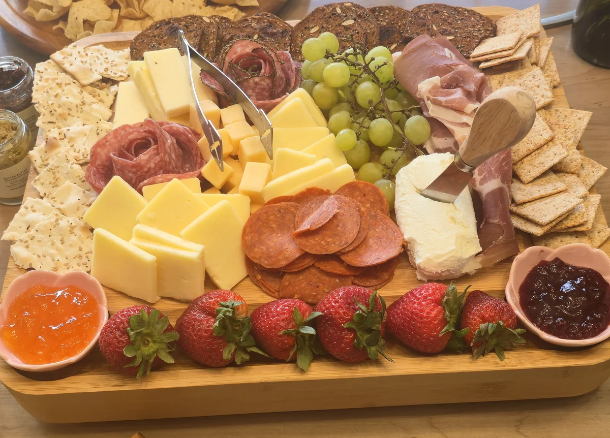 A wooden board filled with strawberries, cheeses, crackers, grapes, meats and preserves